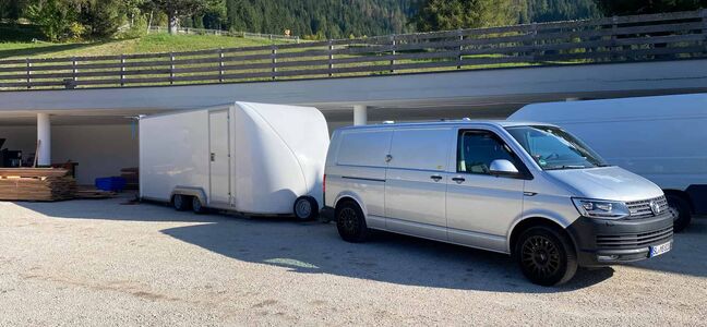 Ein grauer VW Bus mit einem weißen großen Anhänger steht auf einem tiefergelegenen Sandparkplatz. Darüber führt ein Weg oder Straße mit Holzgeländer.